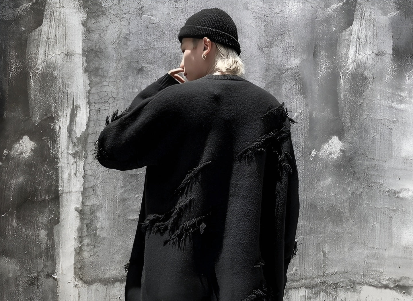 Person wearing an oversized black distressed sweater and black beanie, standing against a rough gray concrete wall with a grunge aesthetic.