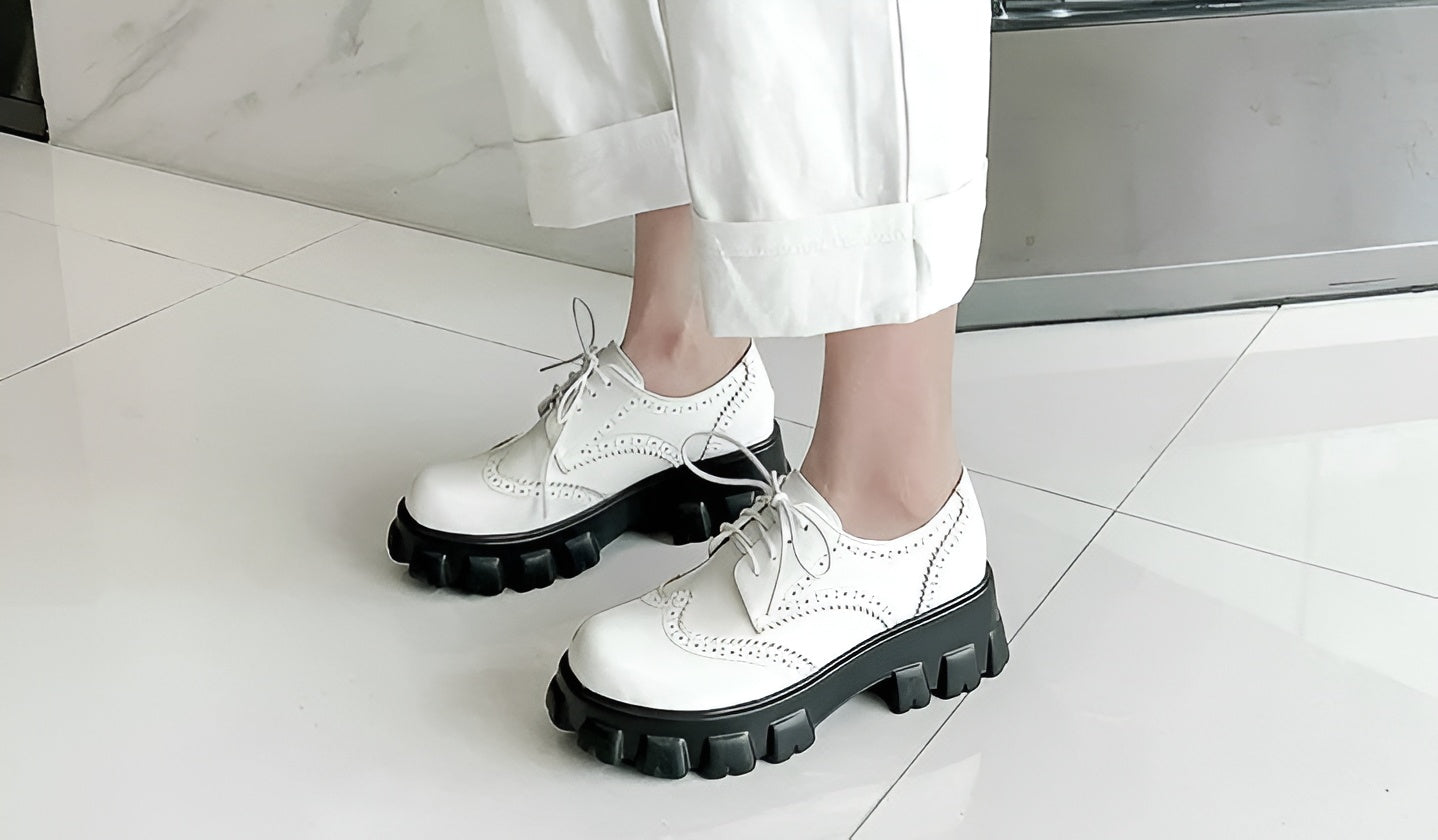White platform Oxford shoes with black chunky soles, worn by a model with white trousers and a black top, showcasing the lace-up design and perforated details.