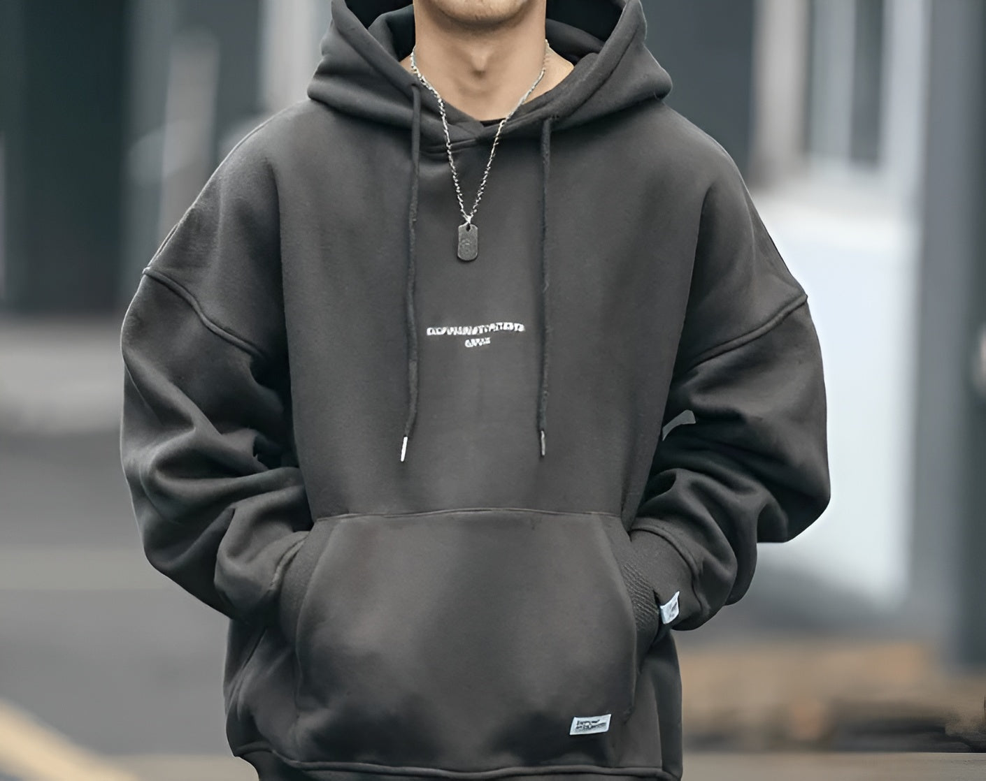 Black oversized men’s hoodie with a small white chest print, worn with a silver chain necklace; casual streetwear style.