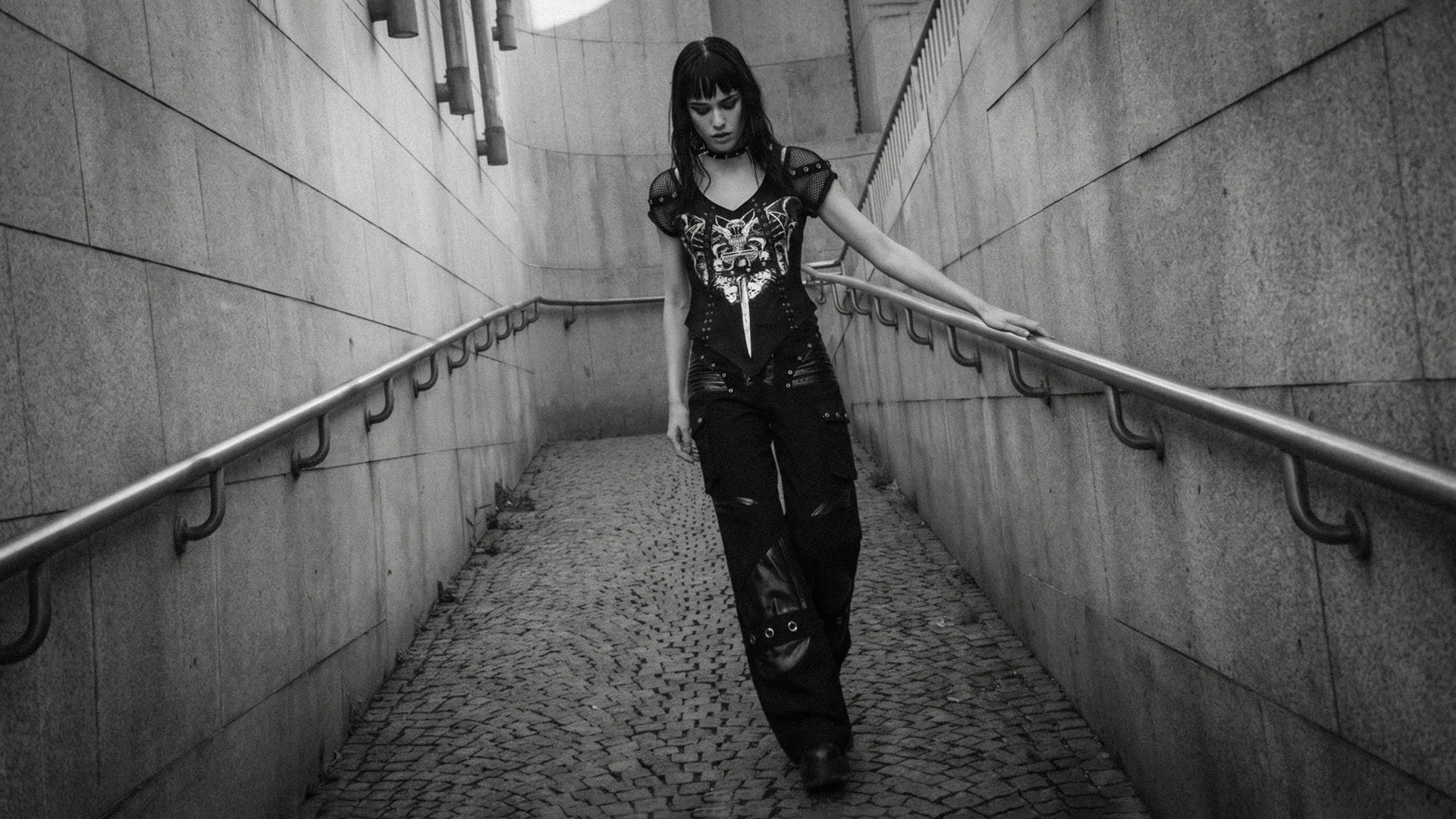 Young woman in a black gothic graphic tee and wide-leg trousers walking down a concrete urban ramp.