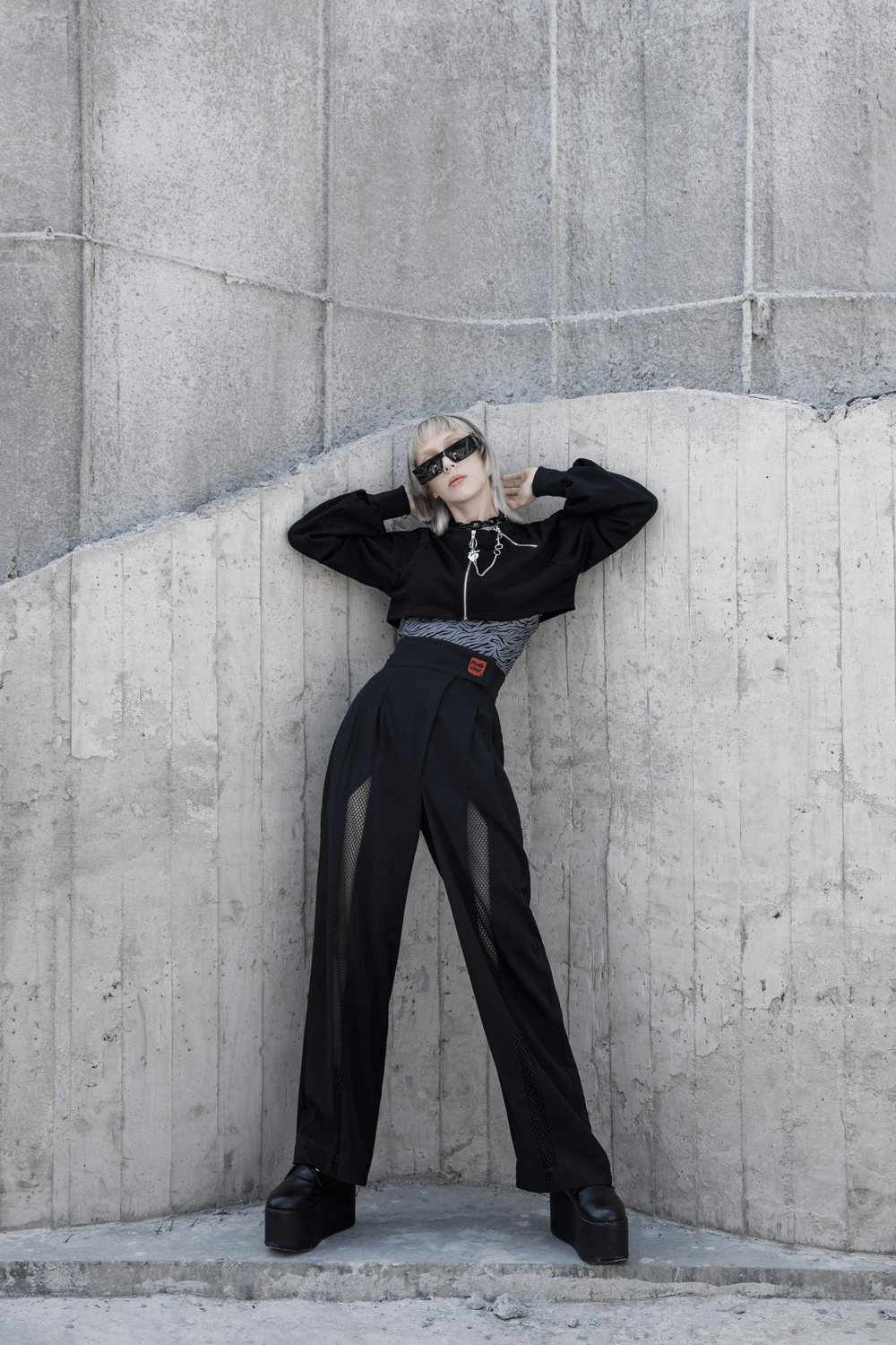 Model in avant-garde black high-waisted wide-leg pants with mesh accents, striking a bold pose against a concrete backdrop.
