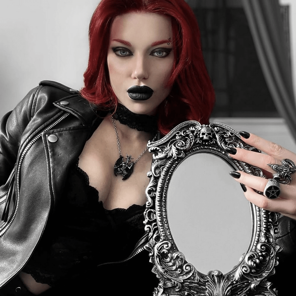 Edgy model with red hair wearing a Baphomet pendant, holding a skull-decorated mirror in a gothic style.