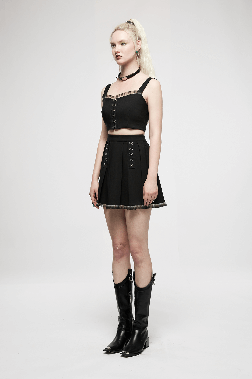 Chic model wearing a black A-line pleated skirt with lace detail and buckle, paired with a stylish crop top and boots.