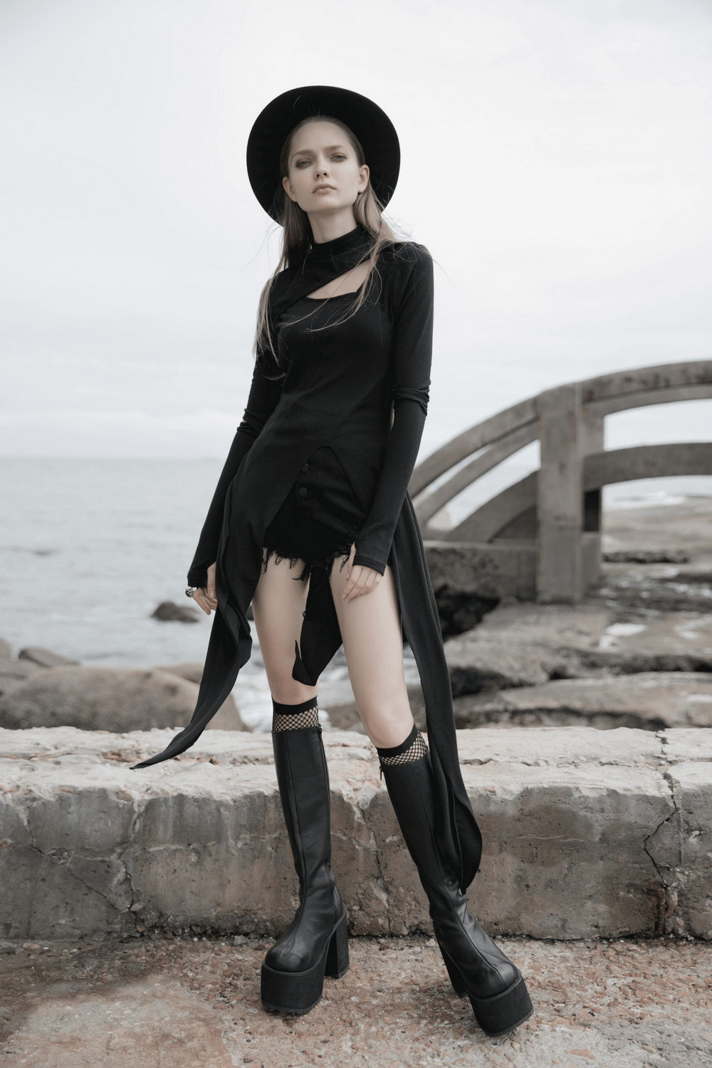 Model wearing a chic black asymmetric knit dress with sleeve cut-outs by the shore, paired with tall black boots and a hat.