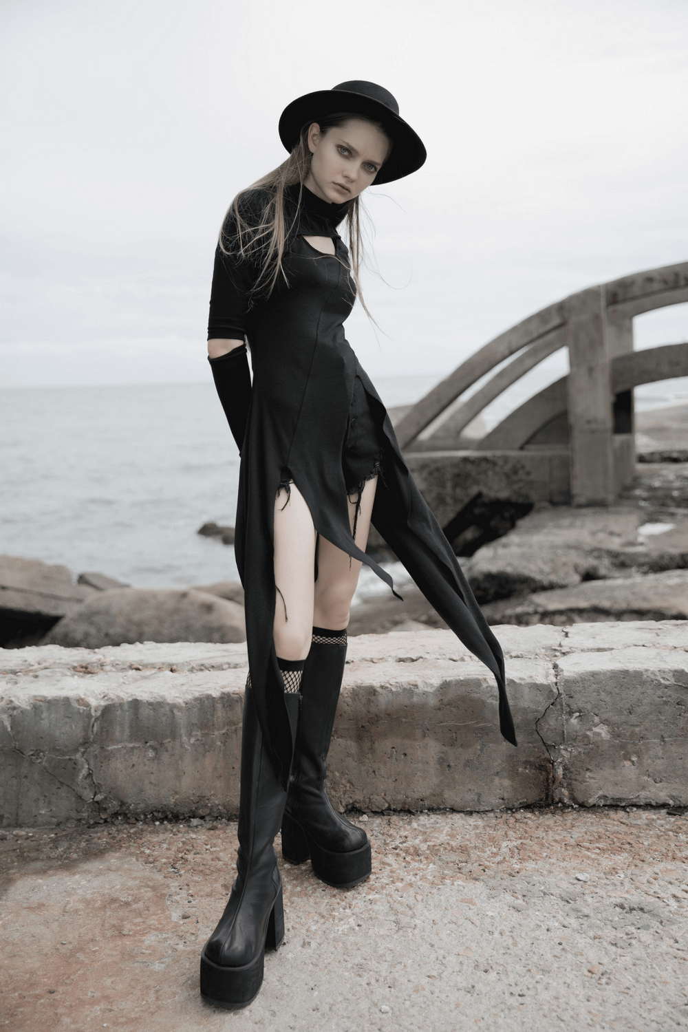 Model wearing a black asymmetric knit dress with sleeve cut-outs, posing by a rocky shoreline in stylish black boots.
