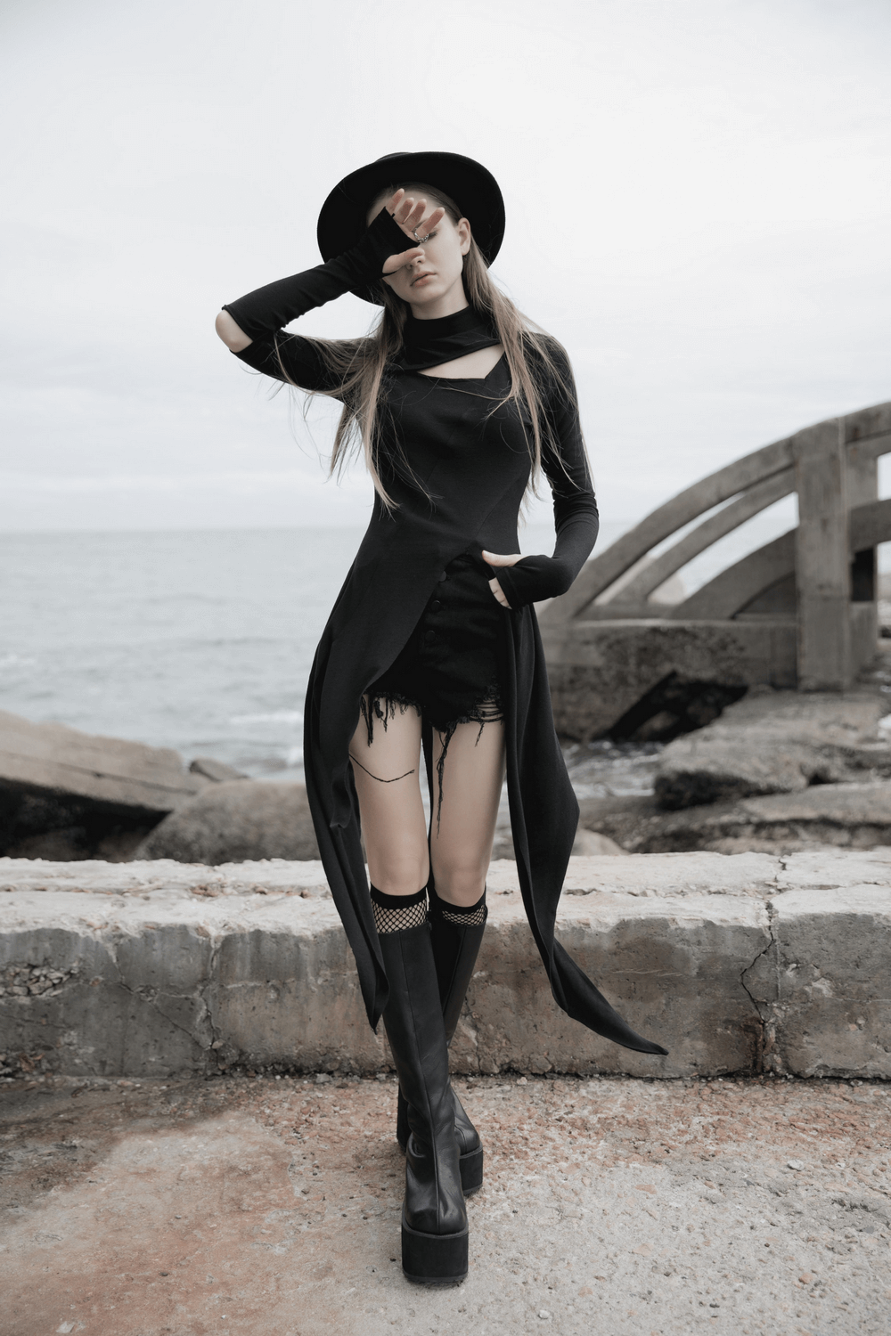 Chic woman in a black asymmetric knit dress with sleeve cut-outs and a hat, posing by the ocean. Fashion-forward style.