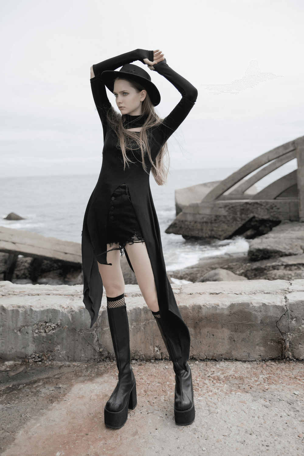 Model showcasing a black asymmetric knit dress with sleeve cut-outs and stylish tall boots on a rocky seascape.
