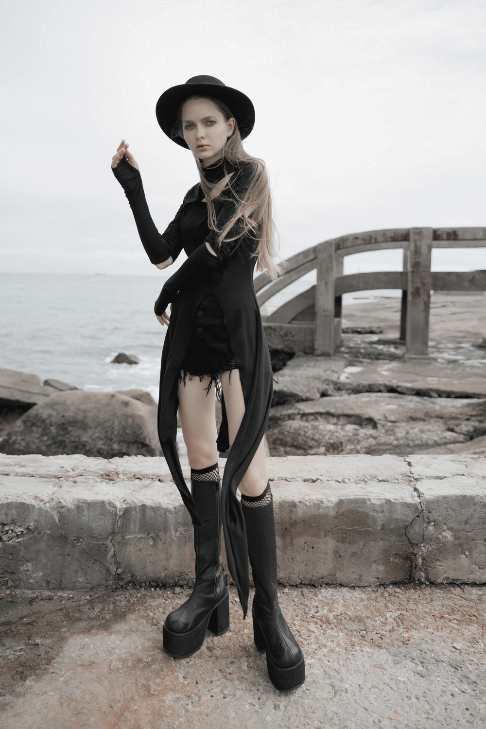 Fashionable model in a black asymmetric knit dress with sleeve cut-outs, standing by the sea.
