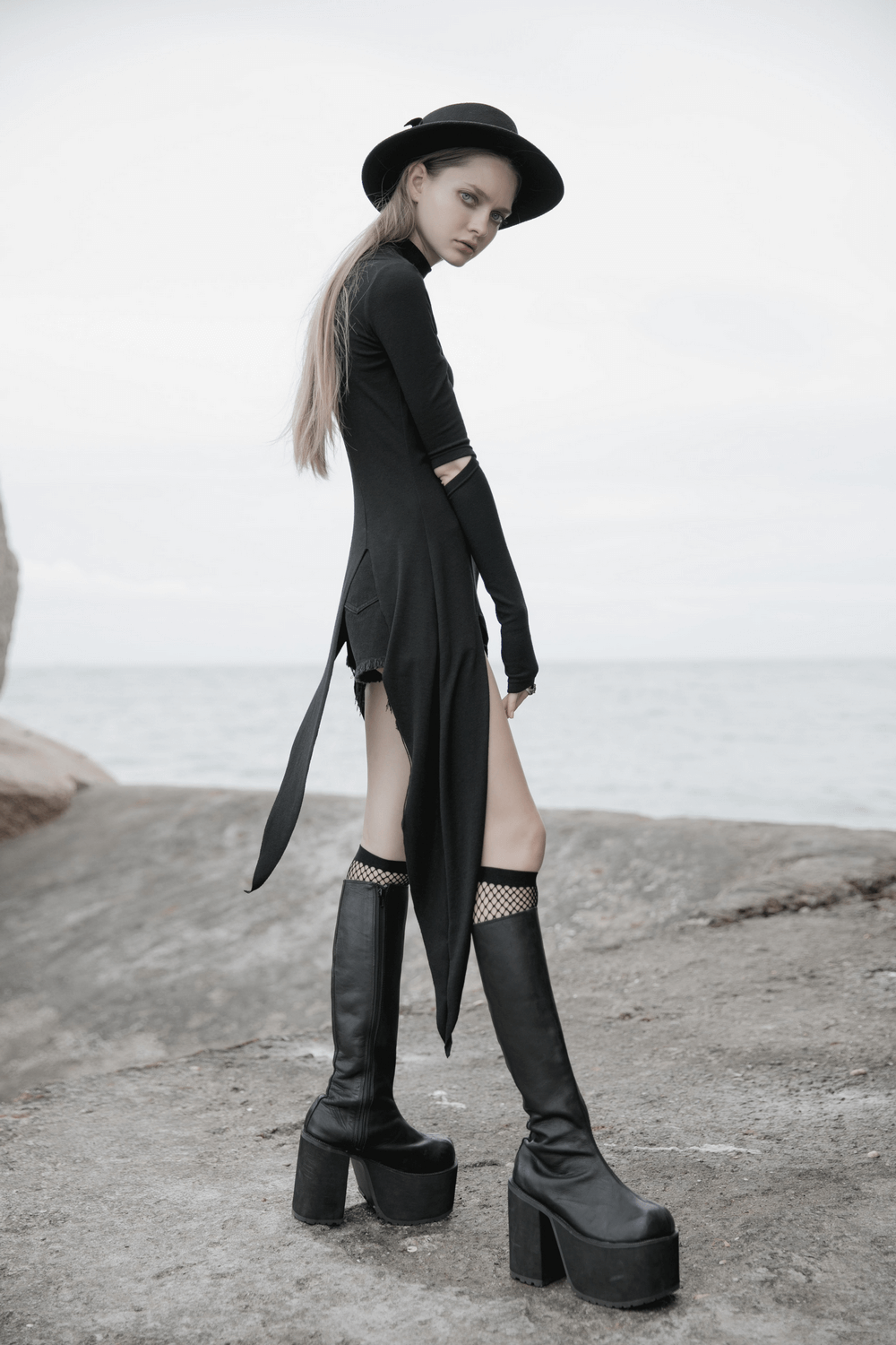 Model showcasing a chic black asymmetric knit dress with sleeve cut-outs, paired with stylish knee-high platform boots.