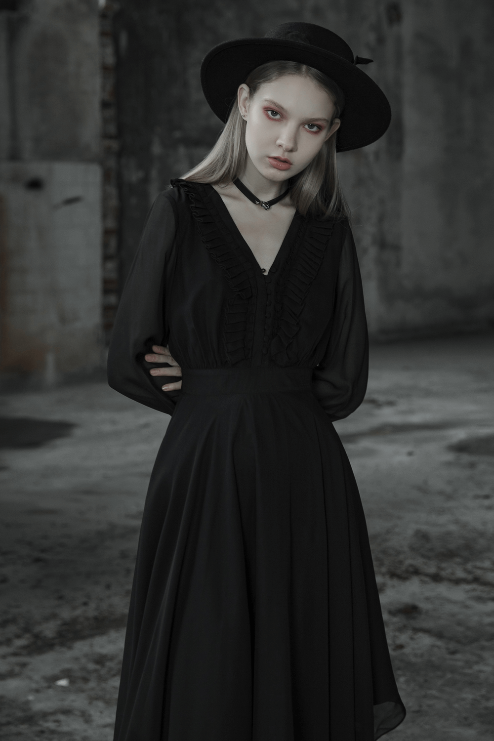 Woman in a black chiffon dress with asymmetrical hem and a stylish hat, posing in a moody urban setting.