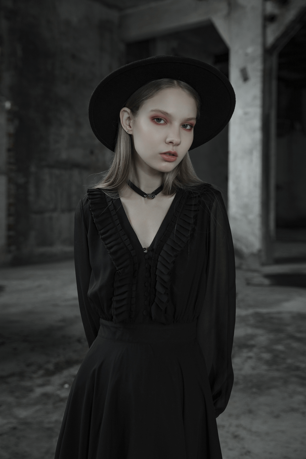 Elegant woman in black dress and hat, showcasing gothic style with ruffled details and striking makeup in an atmospheric setting.