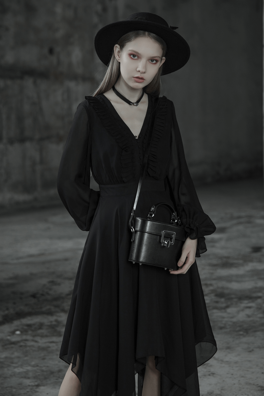 Elegant woman in a black chiffon dress with asymmetrical hem, black hat, and stylish bag, exuding gothic charm.