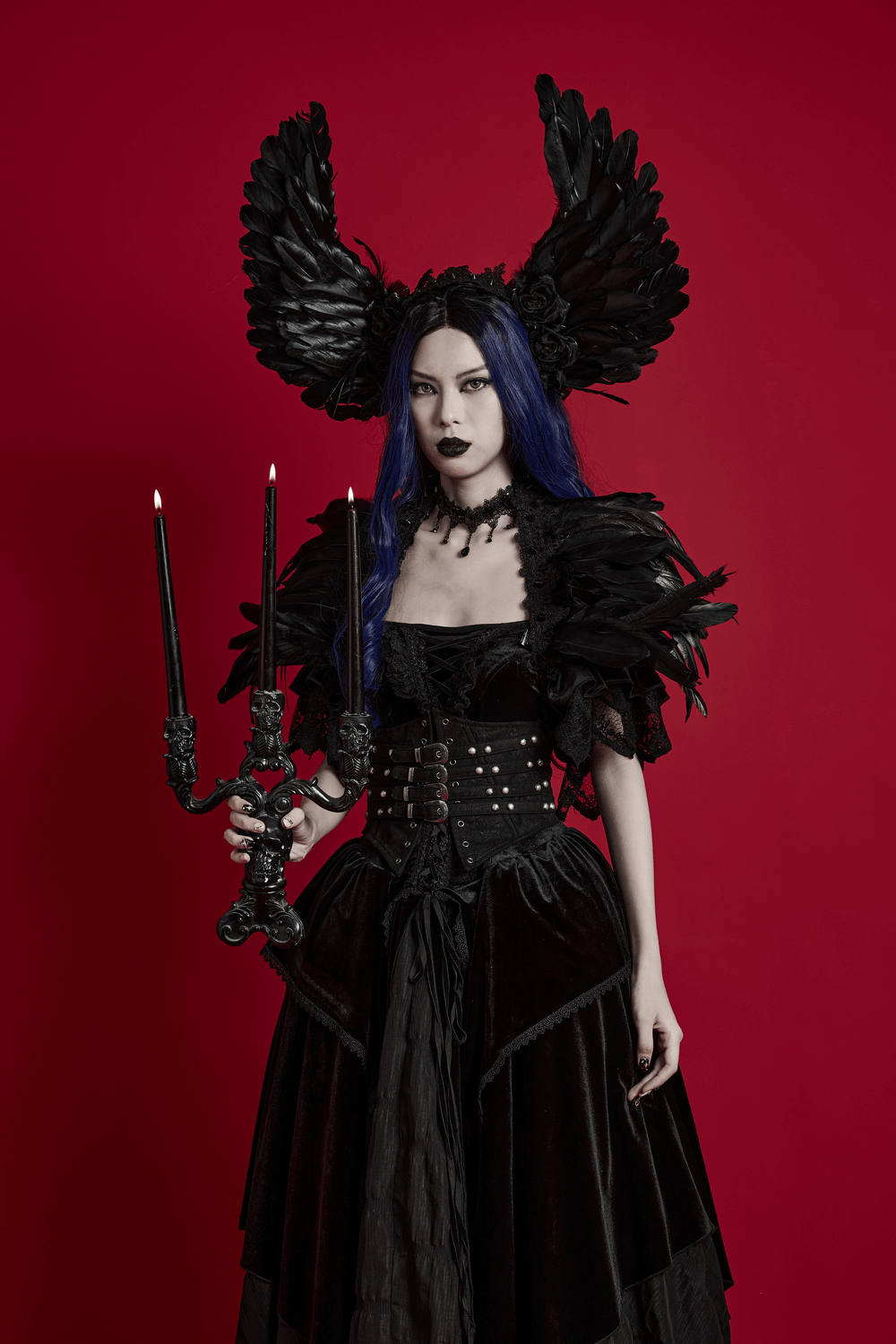 Gothic model in black feather angel wings headband, holding candle holder, wearing dark elegant attire against red background.