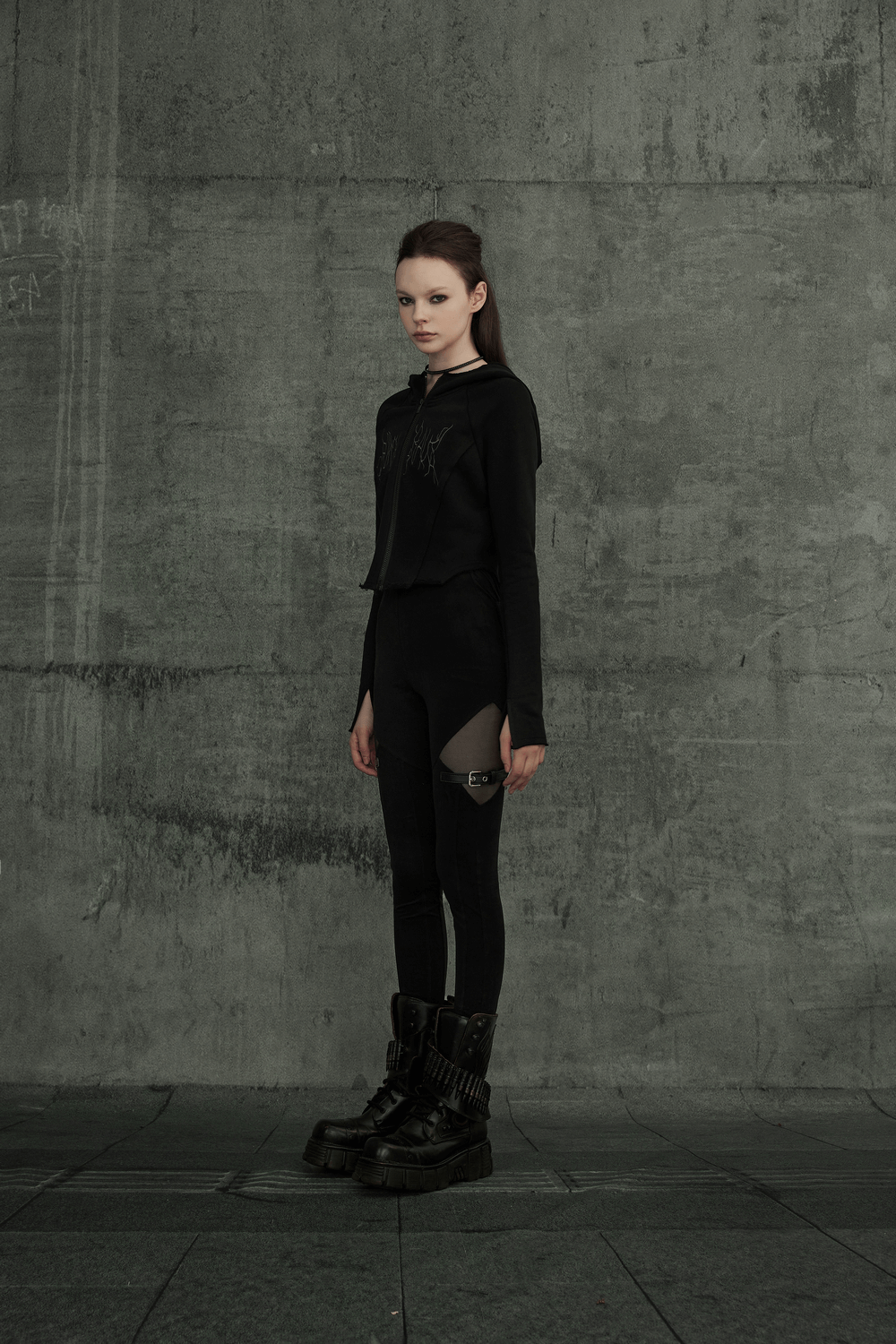 Model in black gothic mesh-panel leggings with buckle accents, exuding edgy punk style against a textured gray backdrop.