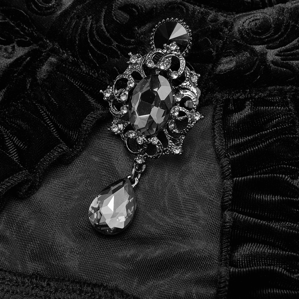 Ornate gothic gem brooch detail on black velvet fabric, featuring intricate designs and a sparkling teardrop pendant.