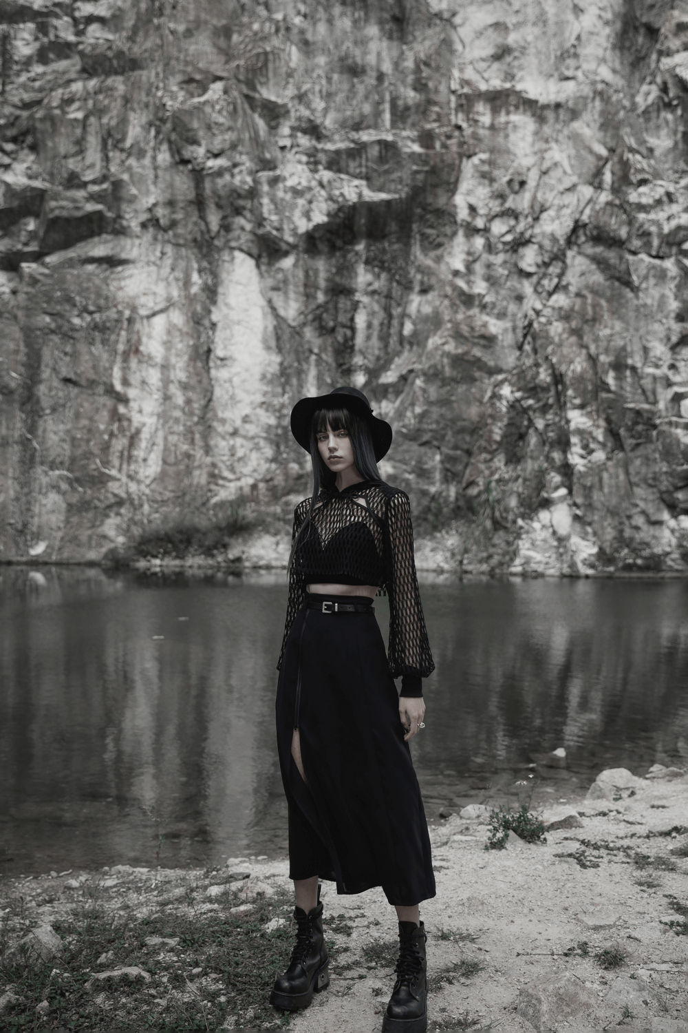 Trendy model wearing a black sheer mesh crop top and hat by a serene lake, showcasing punk rave-inspired fashion.
