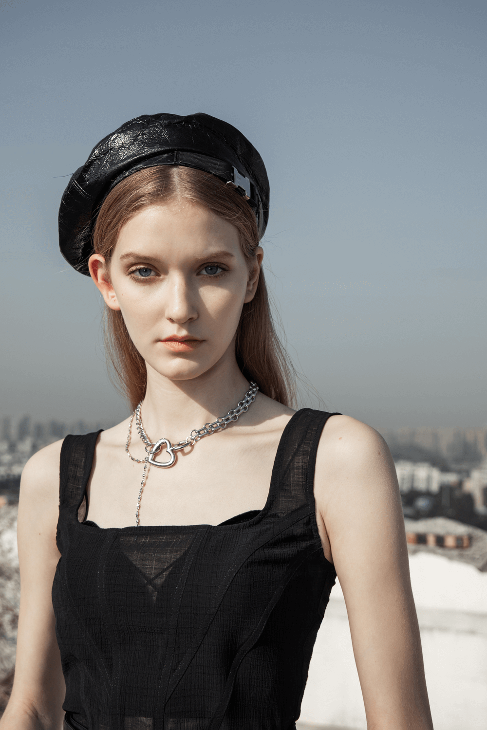 Model wearing a chic black outfit with a stylish beret and heart-shaped necklace against an urban skyline.