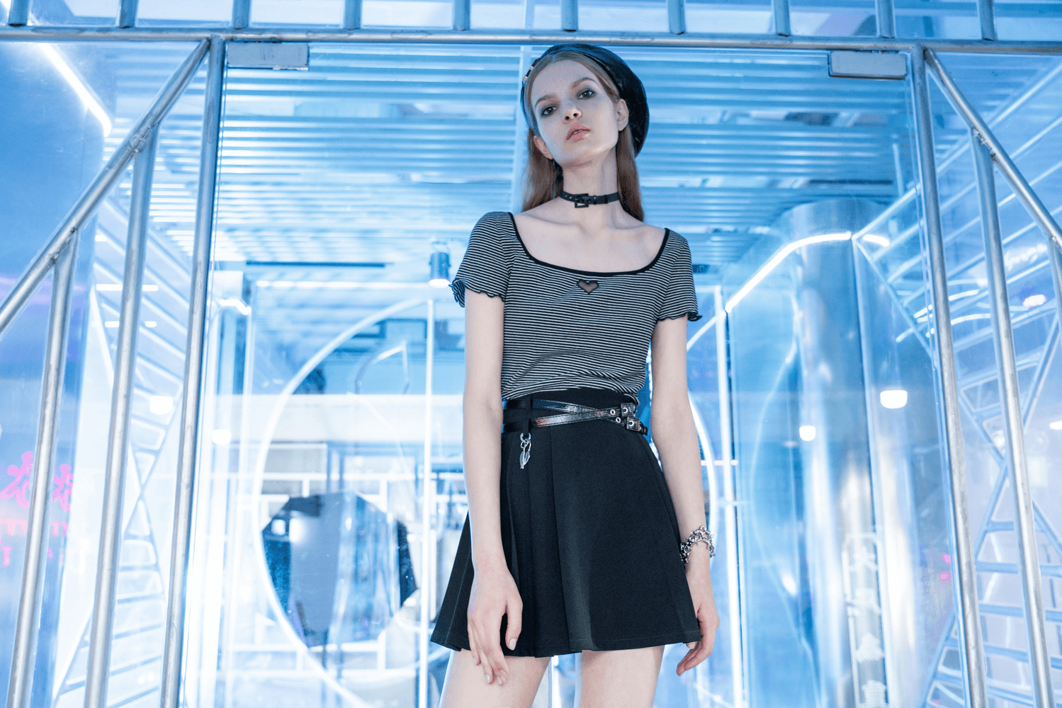 Chic model wearing grey top and pleated black skirt with heart pendant, set against a modern blue-lit background.