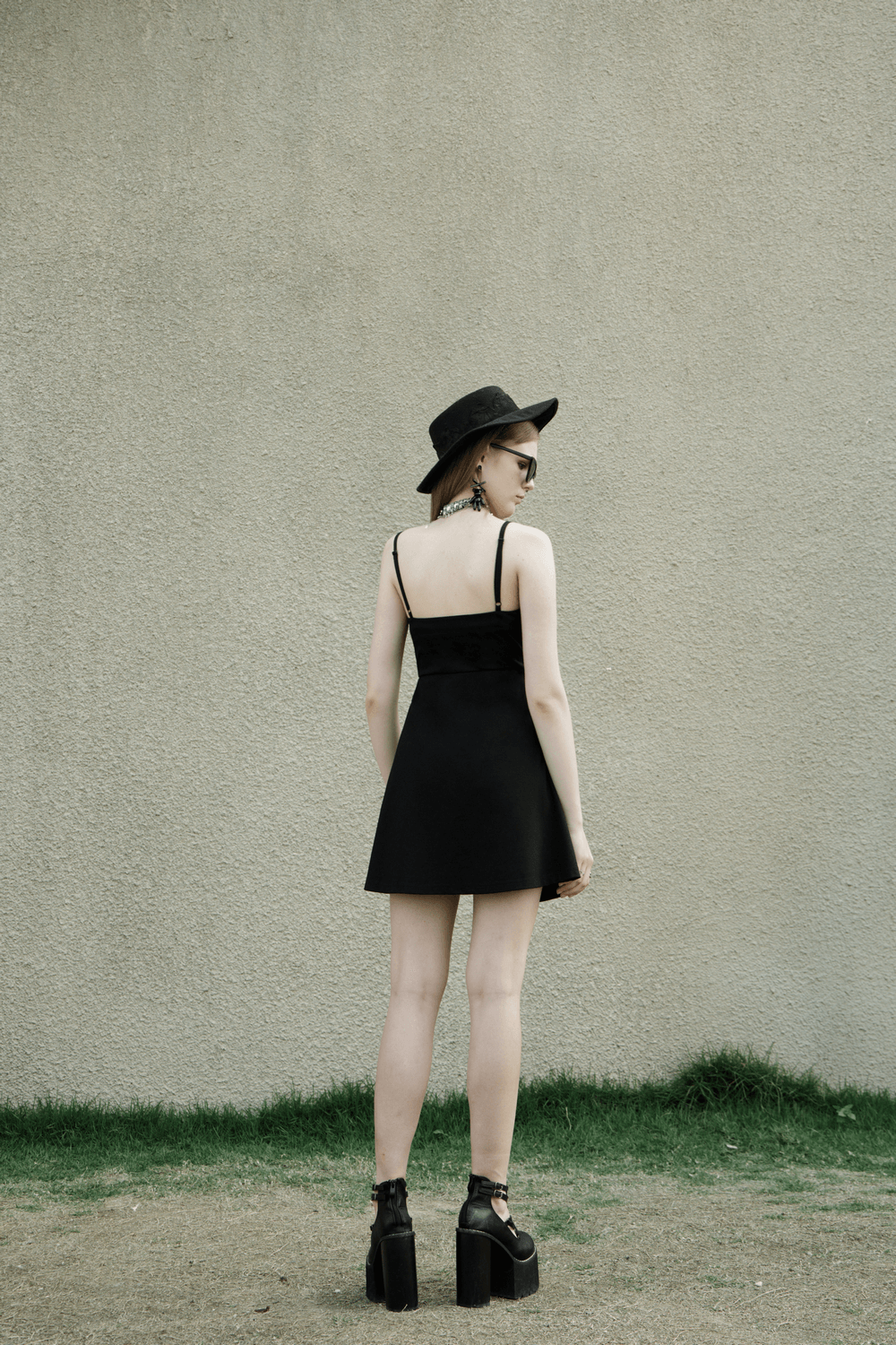 Back view of a chic black V-neck dress with adjustable straps, paired with a stylish hat and platform shoes.