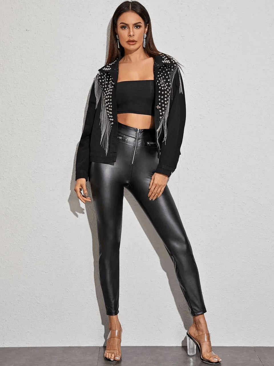 Stylish woman in black denim jacket with tassels and rivets, paired with sleek leather pants and heels for chic streetwear.