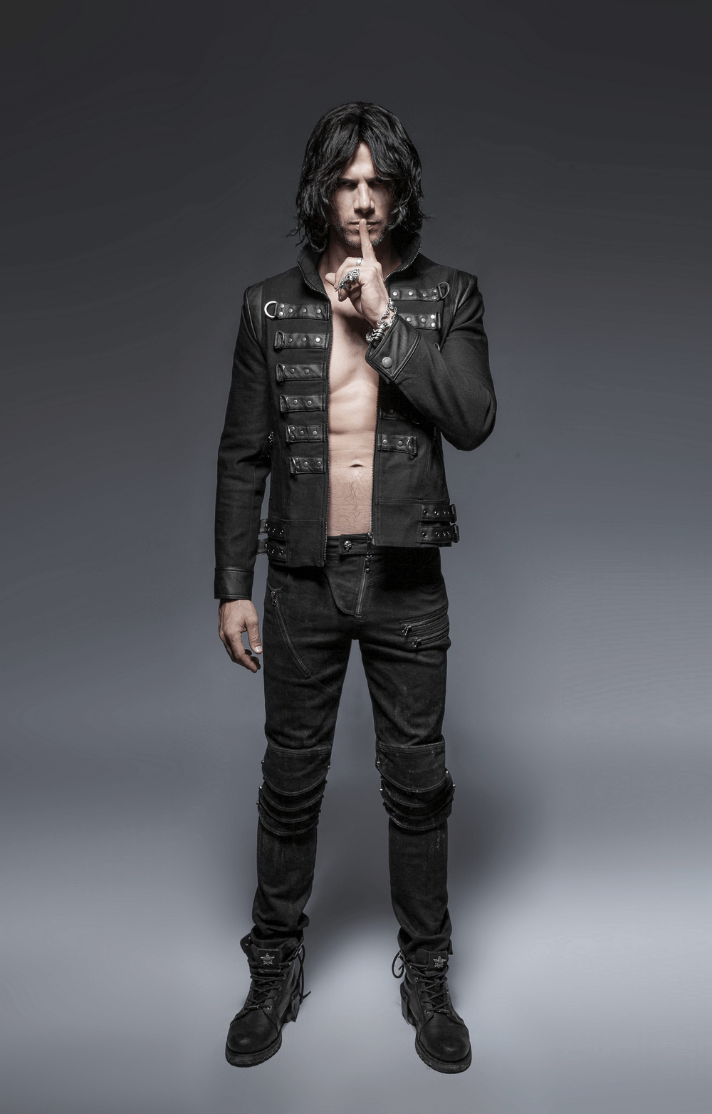 Edgy model in distressed black denim jeans and punk jacket, striking a bold pose with a finger on lips.