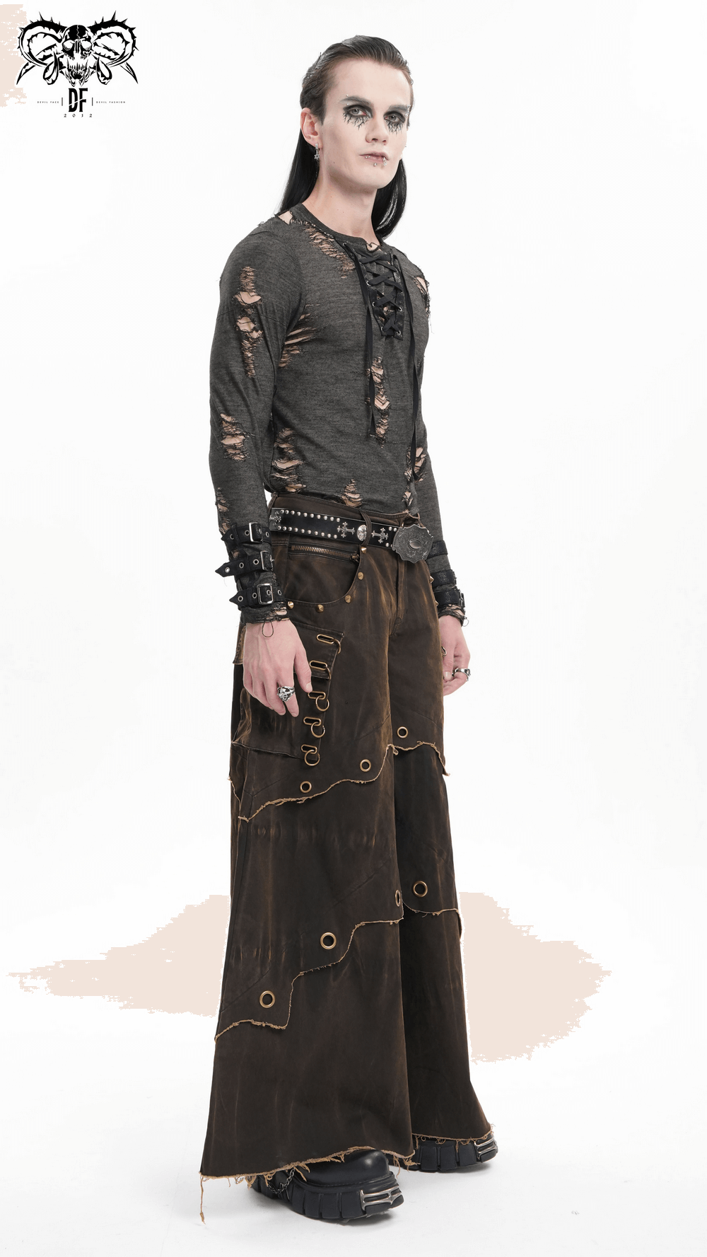 Male model in distressed wide leg pants with grommets, wearing a black layered top and edgy accessories.