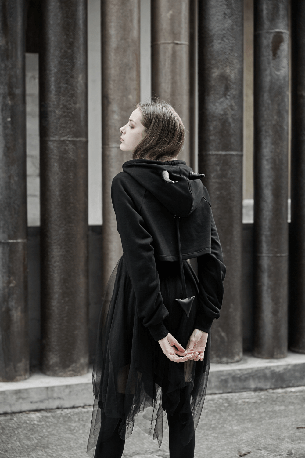 Edgy black crop hoodie with devil horns and detachable tail, stylishly worn against a striking urban backdrop.