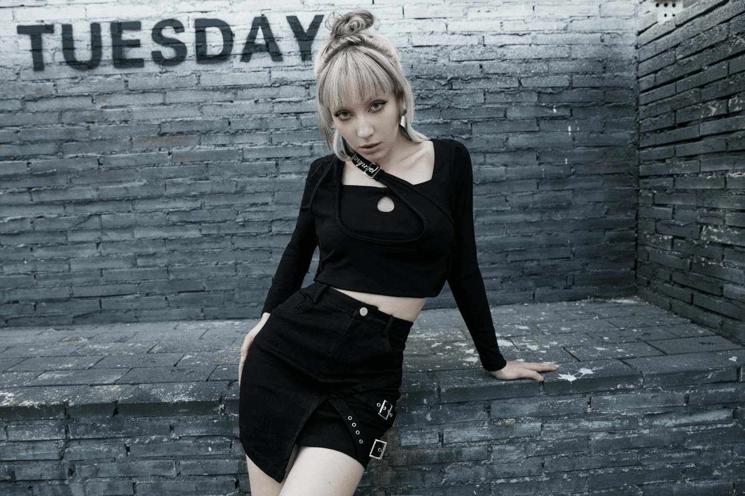 Edgy black denim mini skirt with buckle detail, paired with a chic black crop top against a textured brick backdrop.