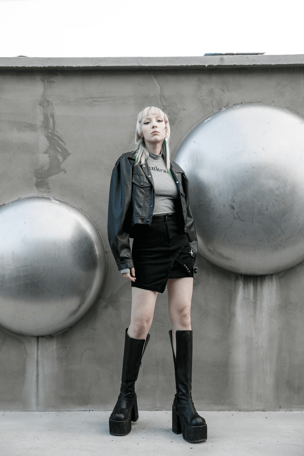 Edgy model wearing a black denim mini skirt with buckle detail and a leather jacket, standing against a bold backdrop.