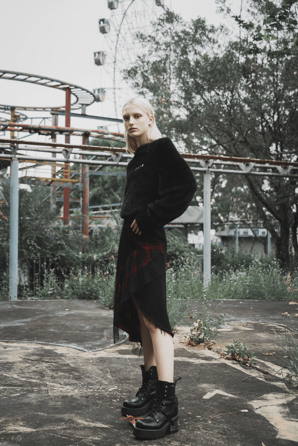 Model wearing an edgy high-waist plaid skirt with irregular hem and black boots in an abandoned park setting.