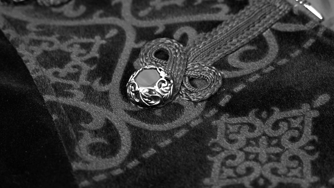 Close-up of a vintage black coat's intricate embroidery and engraved silver button, showcasing gothic elegance.