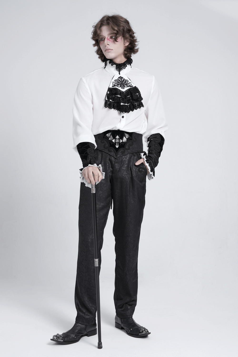 Gothic fashion model in elegant white shirt with lace and black trousers, holding a cane, showcasing vintage aristocratic style.