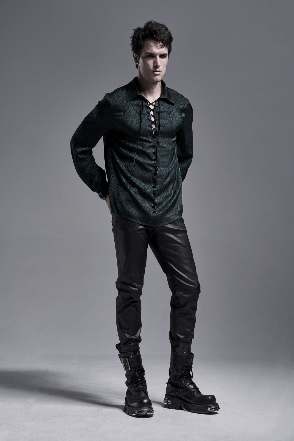 Stylish man in elegant vintage lace-up jacquard shirt with black leather pants and boots on a gray background.