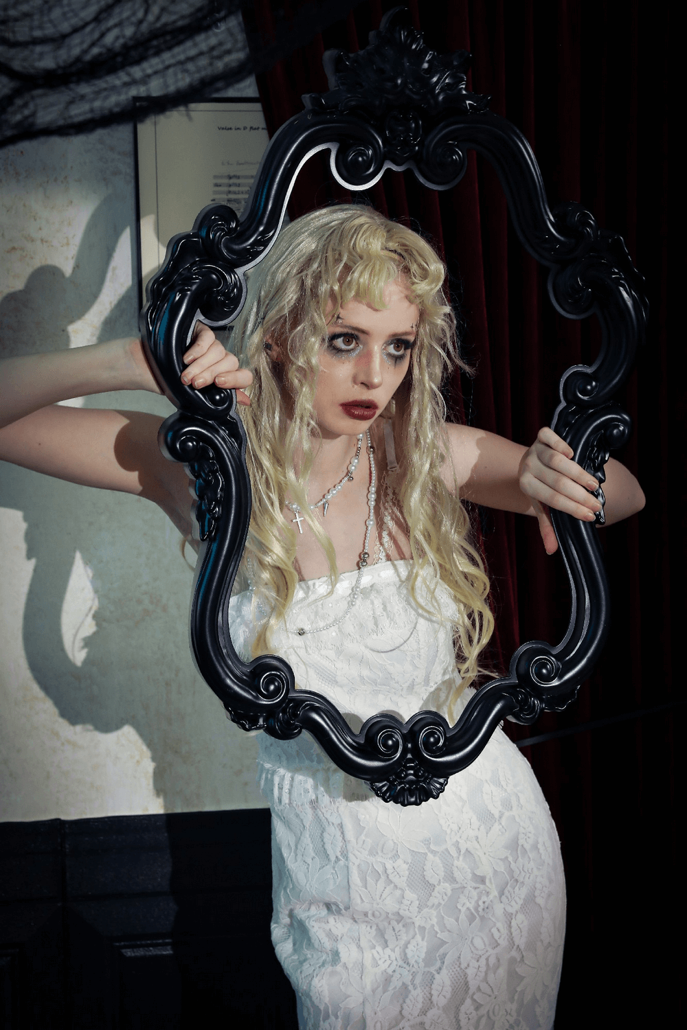 Model in elegant white lace dress holding a black ornate frame in a moody interior setting.