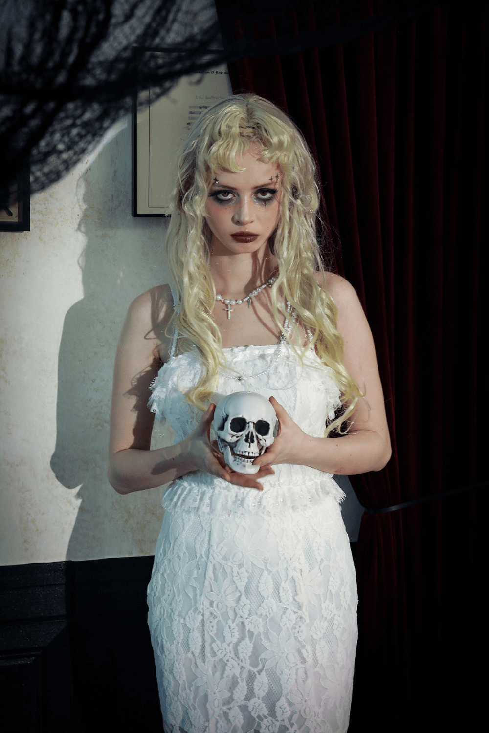 Model in elegant white lace gown holding a skull, set against a dark curtain backdrop, with dramatic lighting effects.