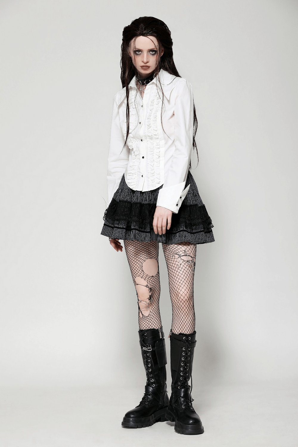 Model in a white ruffled blouse and black mini skirt with fishnet stockings and boots, showcasing edgy chic fashion.