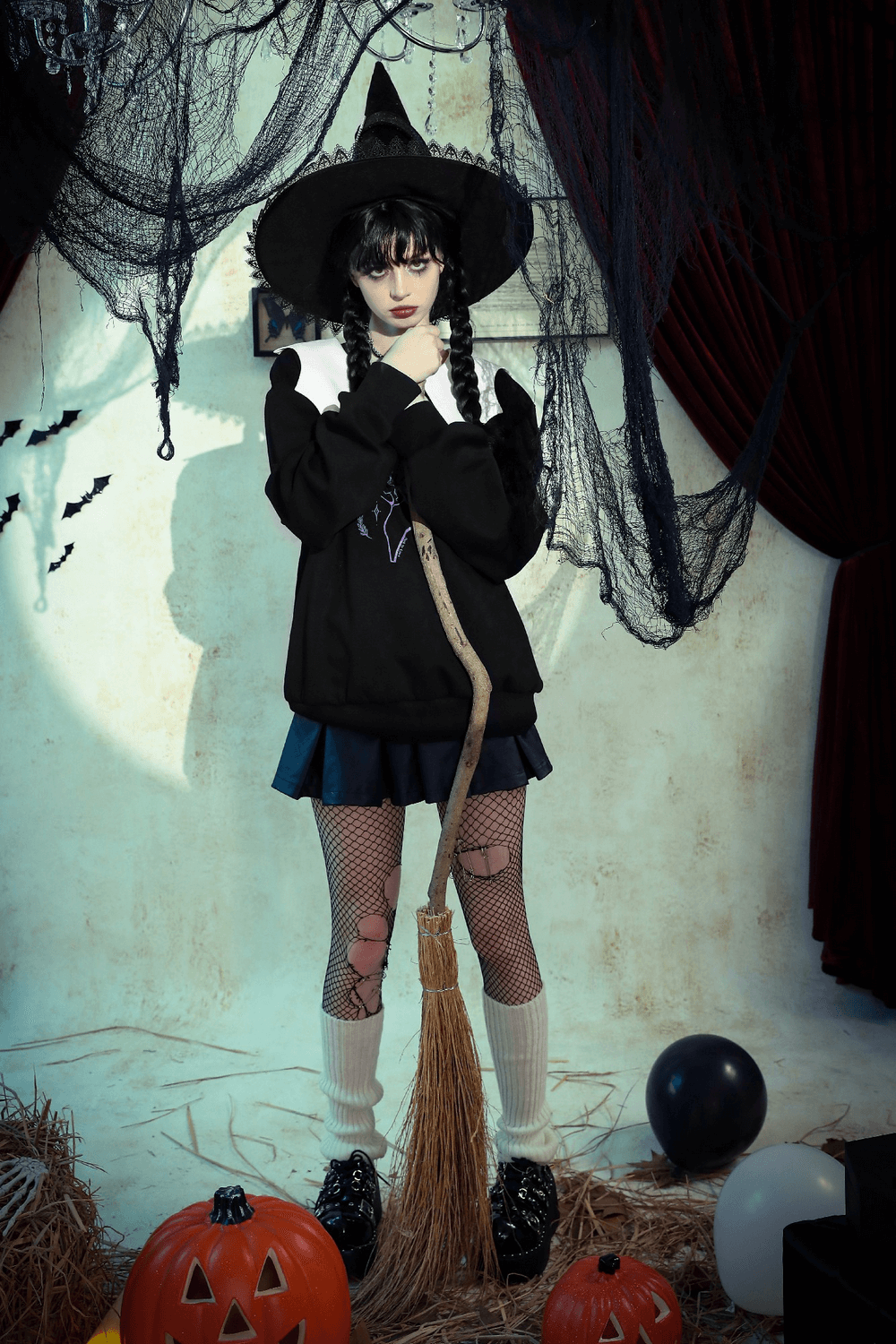 Model wearing a gothic full moon witch sweatshirt with a hat, posing with a broomstick amidst Halloween decorations.