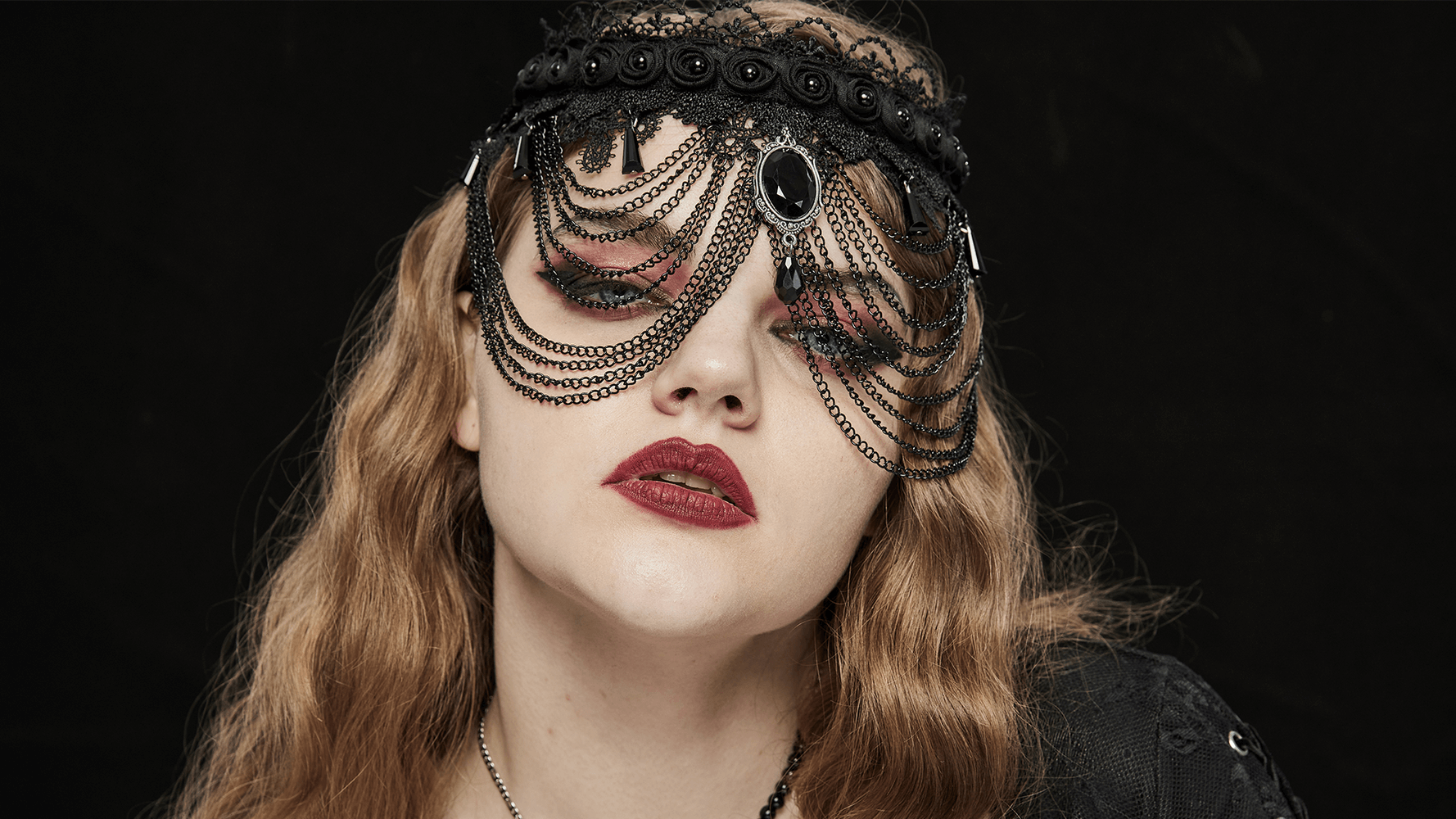 Gothic style woman wearing black lace and chain accessory, showcasing a striking gemstone with bold makeup.