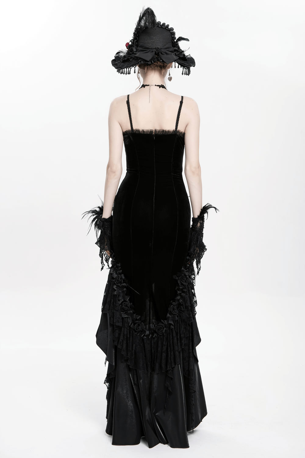 Back view of gothic black velvet high-low evening dress with lace ruffles and satin rose trim, paired with a dramatic hat.