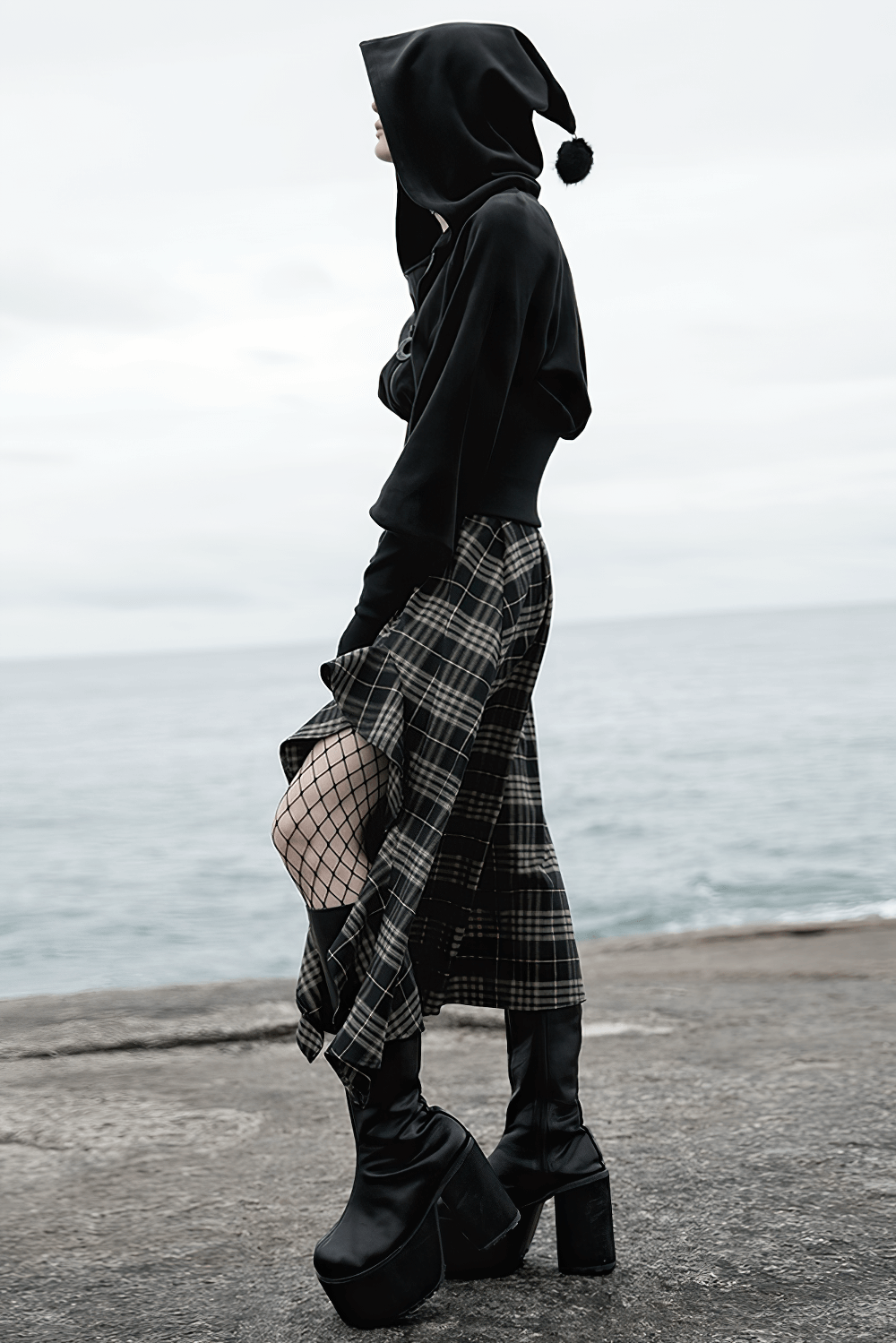 Gothic black velvet hoodie with pointed wizard hood and plaid skirt, capturing edgy fashion by the seaside.