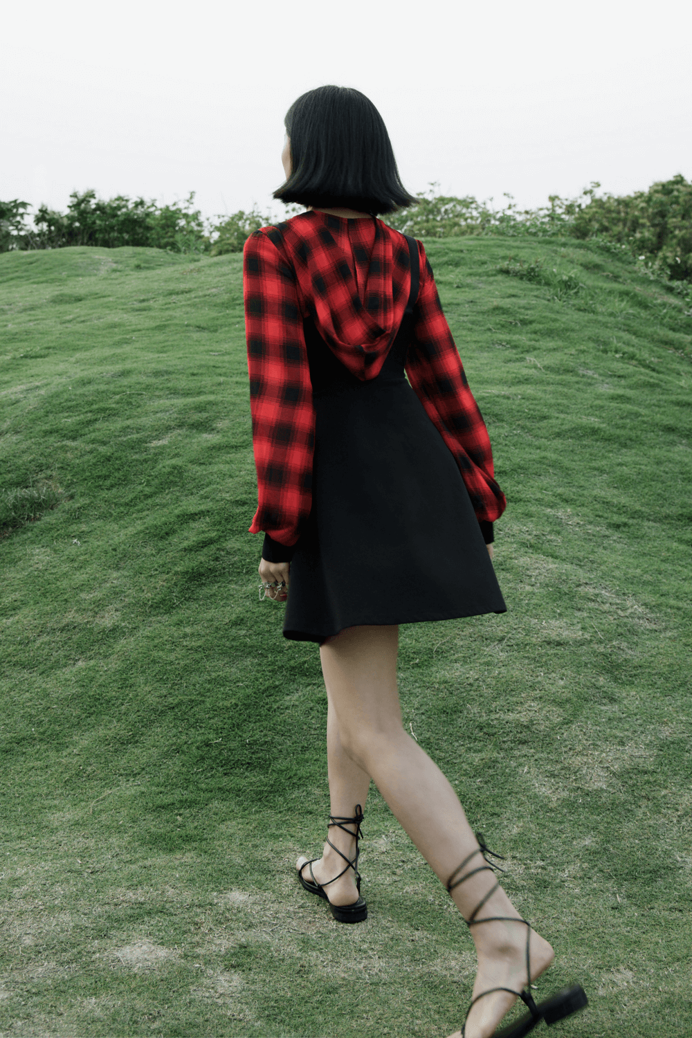 Model walking in Gothic chic plaid hooded mini dress with black bodice and red plaid details on a grassy hill.