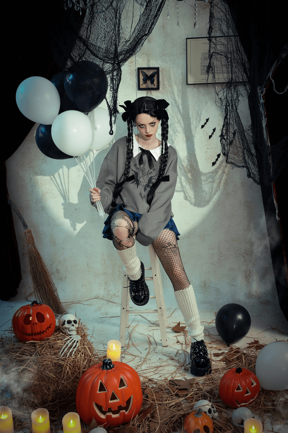 Stylish gothic girl sitting on a stool with black and white balloons, surrounded by pumpkins and Halloween decor.