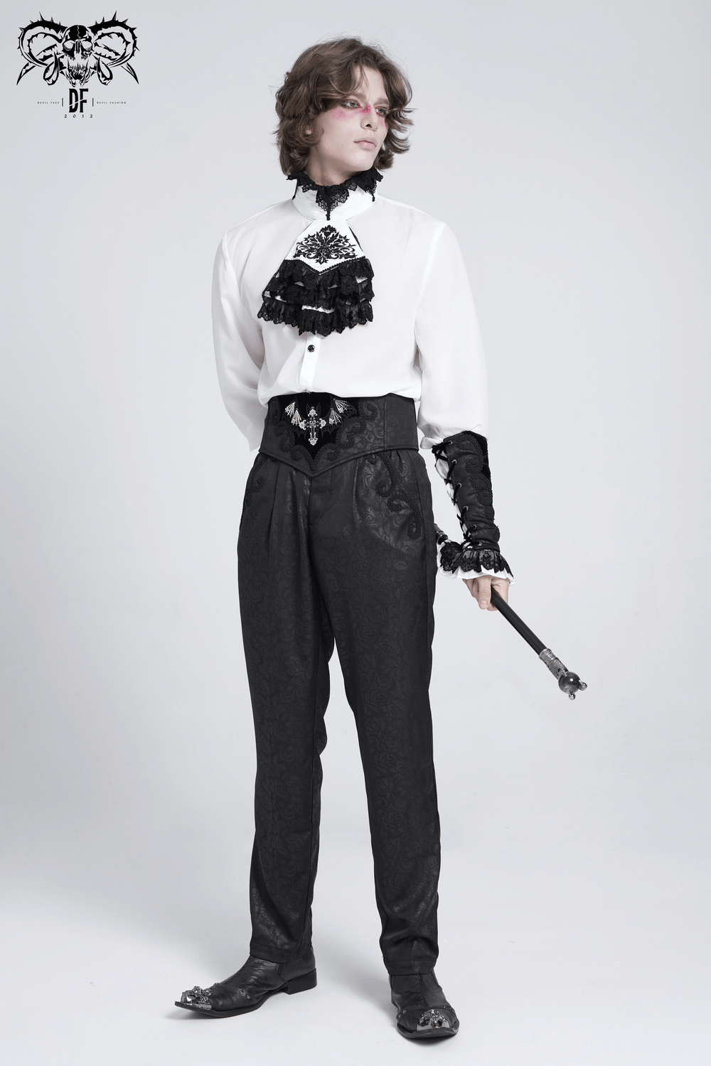 Man in elegant gothic attire with ruffled shirt, high-waisted pants, and cane, showcasing a Victorian-inspired look.