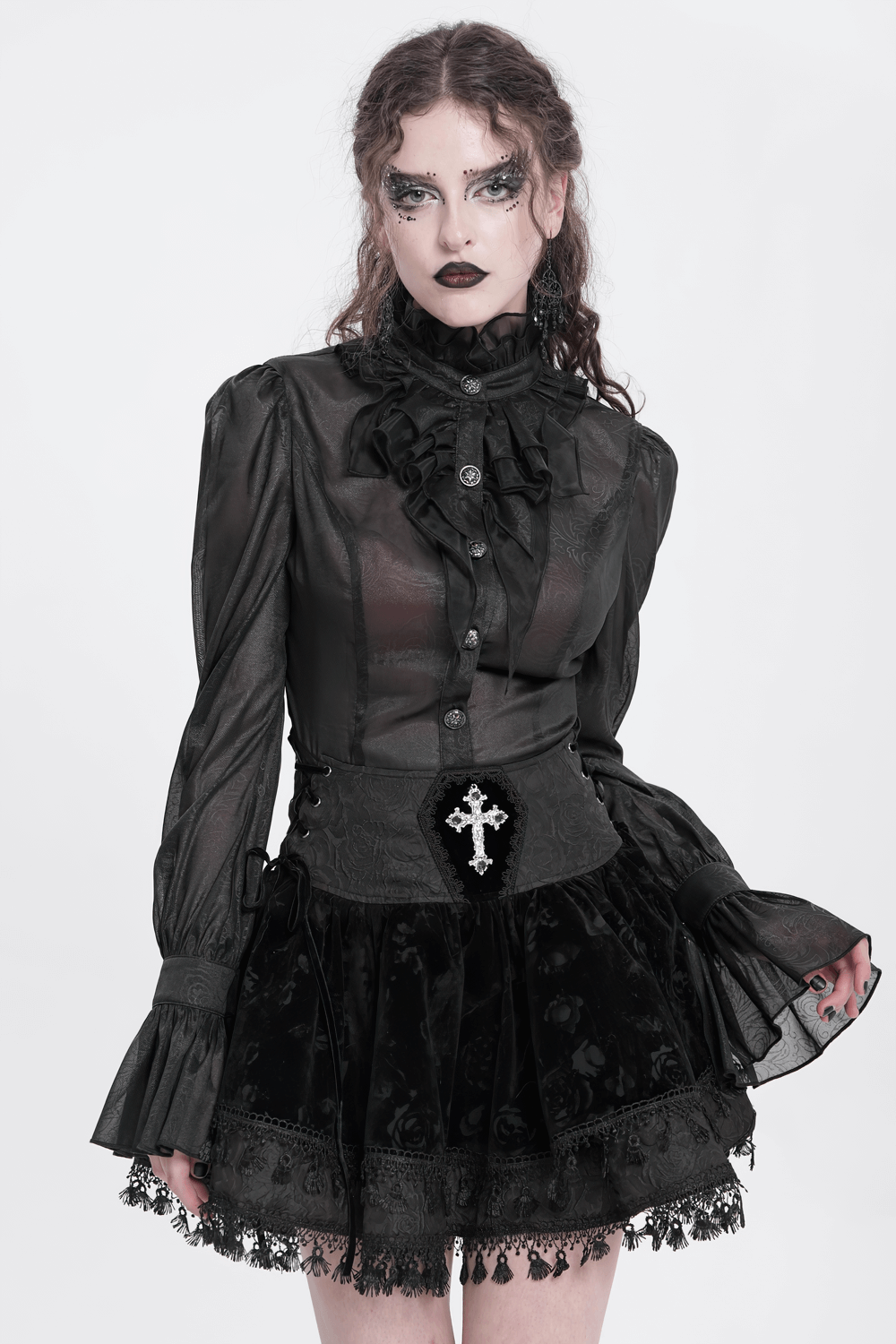 Model in a Gothic lace-up black velvet skirt with embroidered cross and ruffled black blouse against a white background.