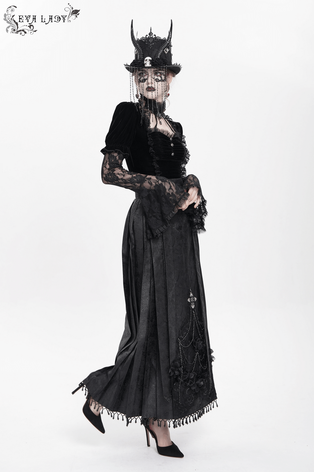 Gothic model in black maxi skirt with lace trim, silver cross, and dramatic hat, showcasing Victorian elegance.
