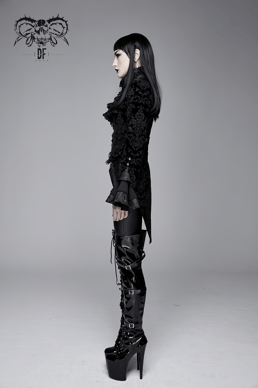 Side view of a woman in a gothic ruffle jacket with lace sleeves and thigh-high boots against a plain background.