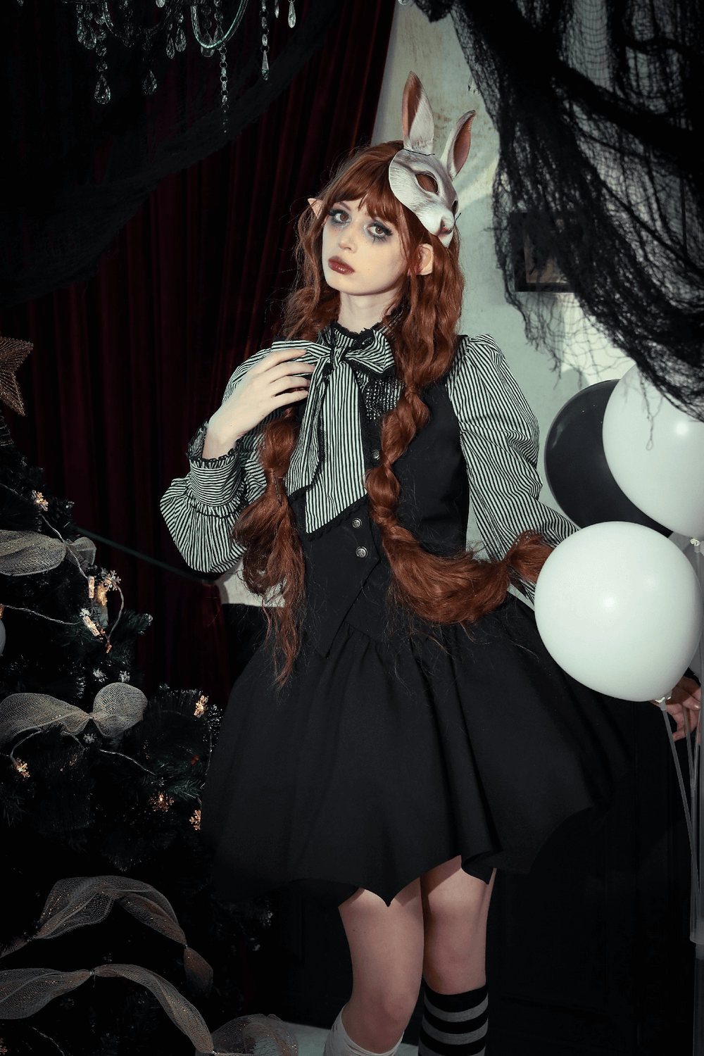 Model in a gothic outfit with bunny mask, black dress, and striped blouse, surrounded by dark decor and balloons.