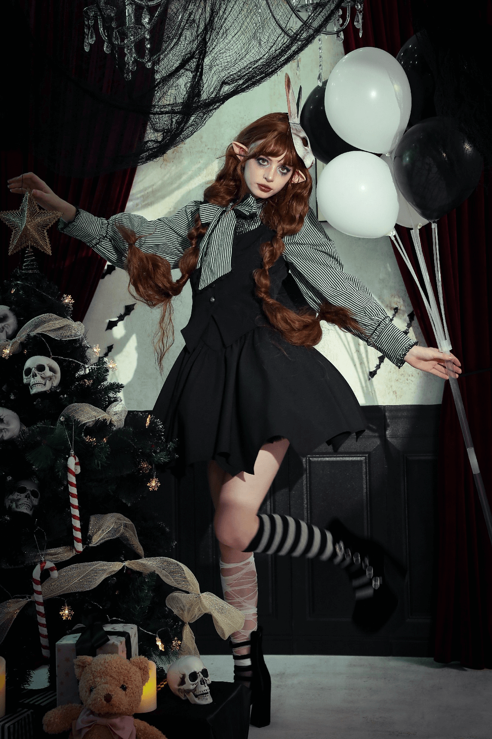 Model wearing gothic outfit with striped socks, surrounded by a decorated Christmas tree and balloons, exuding dark elegance.