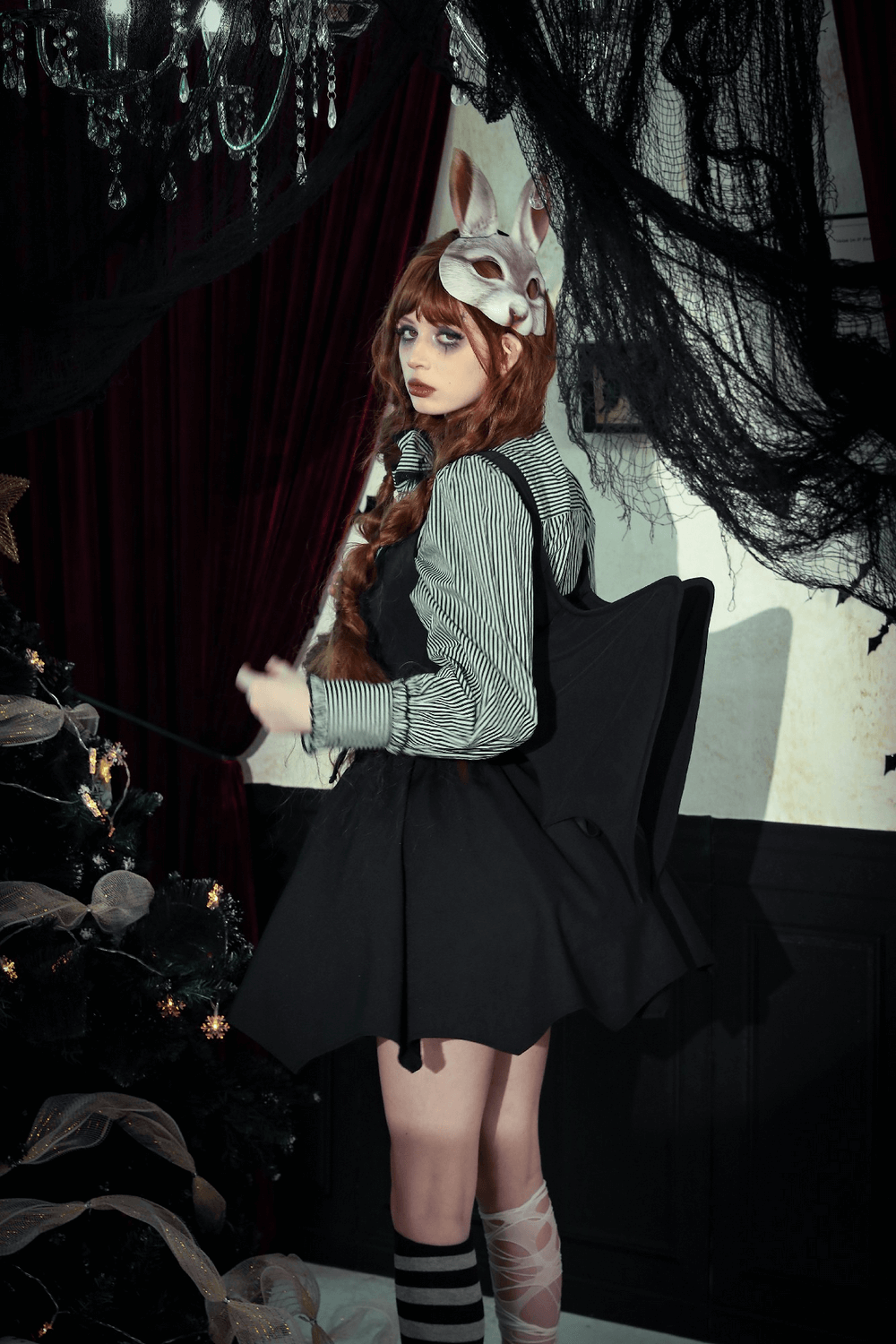 A model in a gothic outfit with bat wings and a bunny mask, posing near a decorated tree in a spooky setting.