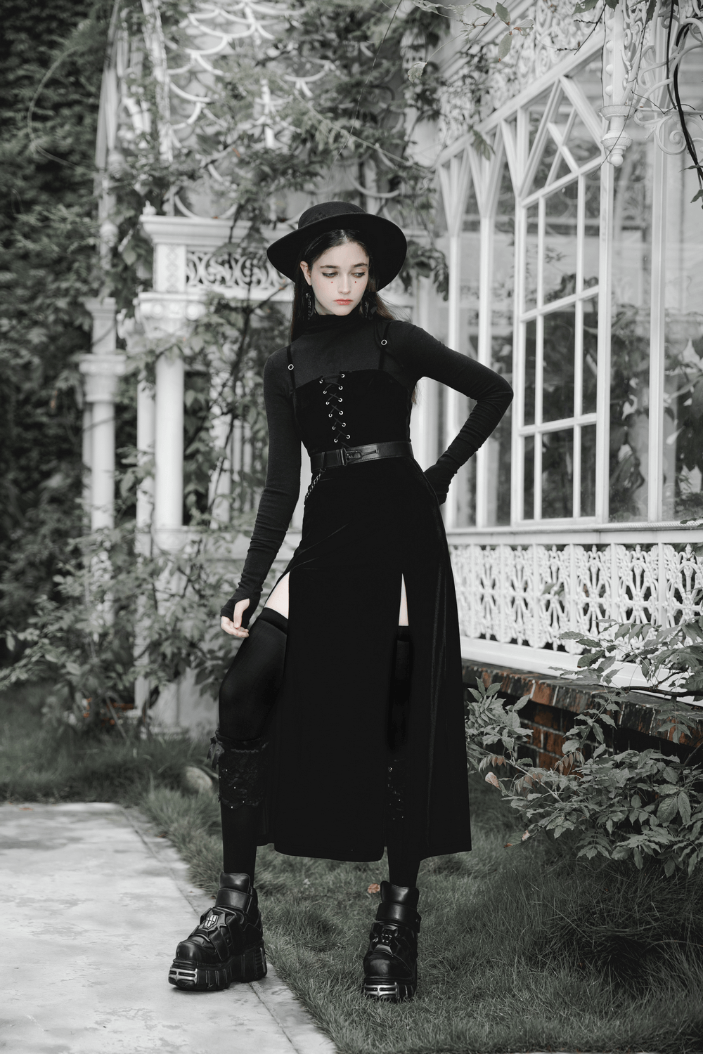 Gothic women's velvet long dress with suspenders and slits, styled with edgy accessories in a garden setting.